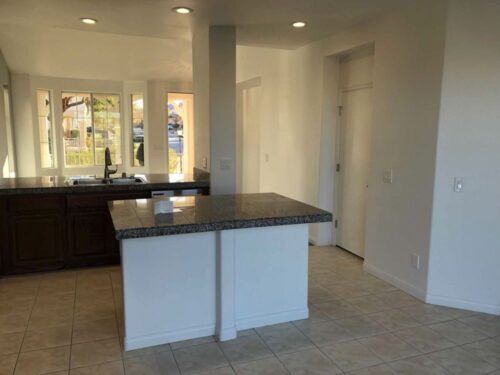 An open white kitchen with gray countertops, recessed lighting and beige tile floors.