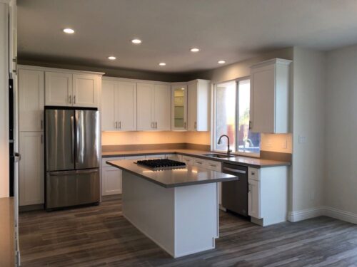 A beige kitchen with white cabinets and gray countertops. Recessed lighting, under cabinet lighting and gray appliances finish it off.