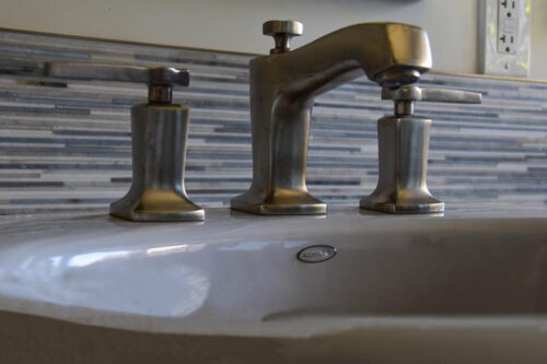 A brushed nickel bathroom faucet with a blue-mixture tile as the backsplash.